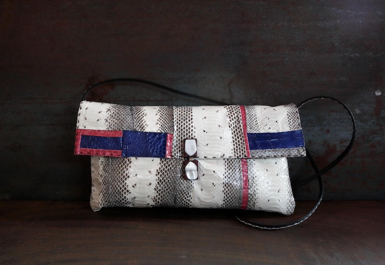 Peut inclure: Pochette rectangulaire avec un motif peau de serpent gris et blanc. Le rabat pr&eacute;sente des accents rouges et bleus. Le sac a une bandouli&egrave;re noire et une fermeture &agrave; fermoir.