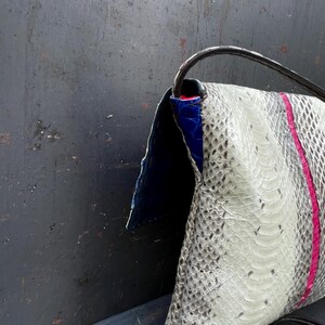 Peut inclure: Un sac pochette avec un motif peau de serpent dans les tons de blanc et de gris, accentu&eacute; d'une bande rose verticale. Le sac pr&eacute;sente un int&eacute;rieur bleu et une bandouli&egrave;re noire. Le sac est fait d'une mati&egrave;re textur&eacute;e.