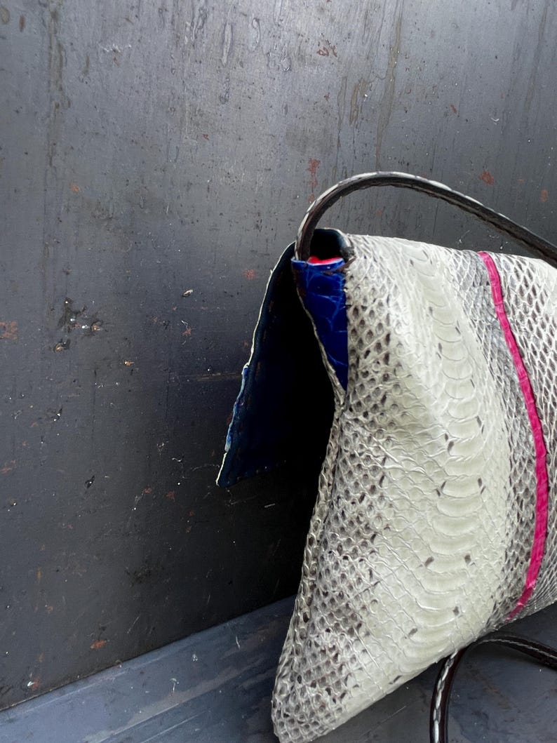 Peut inclure: Un sac pochette avec un motif peau de serpent dans les tons de blanc et de gris, accentu&eacute; d'une bande rose verticale. Le sac pr&eacute;sente un int&eacute;rieur bleu et une bandouli&egrave;re noire. Le sac est fait d'une mati&egrave;re textur&eacute;e.