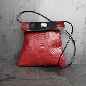 Op de afbeelding: Een rode handtas met slangenleertextuur en een zwarte riem. De tas heeft een zwarte en bordeauxkleurige flap met een zilverkleurige decoratieve sluiting. De tas heeft een vierkante vorm en een lange, dunne zwarte riem.