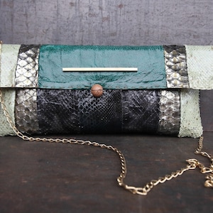 Op de afbeelding: Een groene, zwarte en zilveren slangenleren clutch met een gouden kettingband.