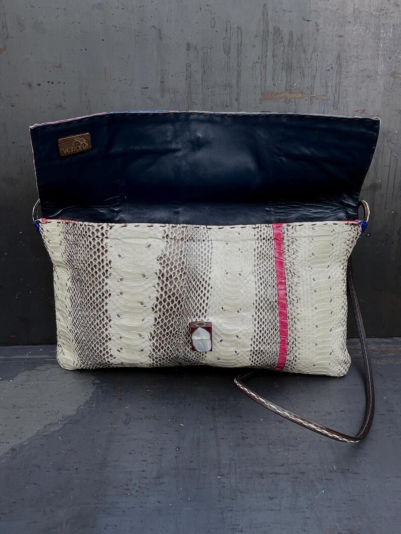 Peut inclure: Pochette rectangulaire avec un motif peau de serpent blanc, gris et marron. Une bande verticale de cuir rose orne le devant. L'int&eacute;rieur est bleu marine. Le sac a une longue et fine bandouli&egrave;re marron.