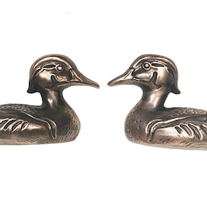 May include: Two bronze-colored duck figurines. Each duck is in a resting position, with detailed feather and beak features. The ducks face each other, creating a symmetrical display. The background is plain white.
