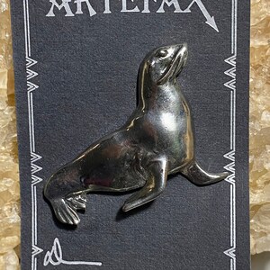 May include: A silver seal pin on a black card with the word "ARTÉFAX" printed in white.