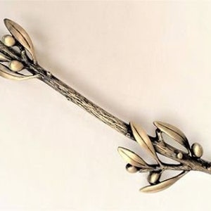 May include: A decorative bronze-toned olive branch with leaves and olives. The branch has a textured, naturalistic appearance, with detailed veining on the leaves. The olives are small and round, adding to the realistic design. The item is likely a home decor accent.