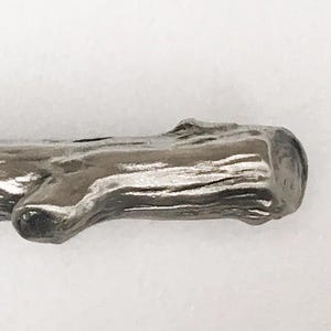 May include: A silver-colored, twig-shaped decorative object. The object has a textured surface, resembling tree bark, with small protruding branches. The ends are rounded, and the overall design is naturalistic.