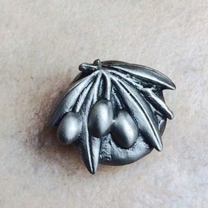 May include: A silver-toned metal pin featuring a stylized olive branch with three olives. The pin is a circular shape and has a detailed design.