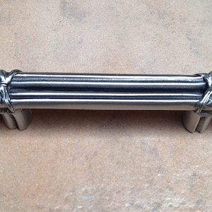 May include: A silver cabinet pull with a design of three stacked, tied, and twisted metal rods.