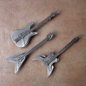May include: Three silver-colored miniature electric guitars. The guitars are in different styles, including a traditional electric guitar, a flying V, and a more modern, angular design.