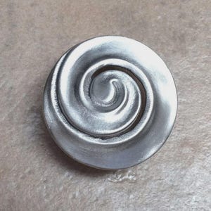 May include: A silver-colored, circular decorative element with a raised spiral design. The spiral is centered within the circle, creating a three-dimensional effect. The surface has a brushed metal texture.