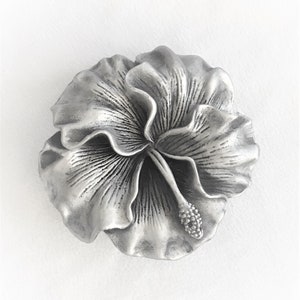 May include: A detailed, silver-colored hibiscus flower sculpture. The flower has multiple layers of petals with textured lines and a central stamen. The sculpture is a decorative piece, likely for home decor or jewelry.