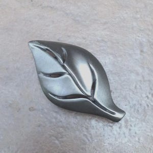 May include: A metallic gray leaf-shaped decorative element. The leaf has detailed veining and a smooth, reflective surface. The design is a stylized representation of a leaf, suitable for home decor or craft projects.