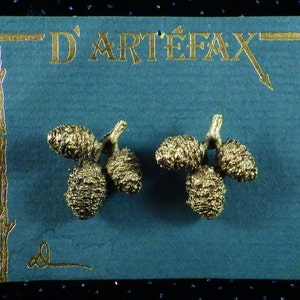 May include: A pair of gold-colored earrings shaped like pine cones, displayed on a teal card with gold lettering that reads "D'ARTEFAX". The earrings are detailed and textured, with a branch-like stem.