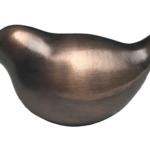 May include: A small, bronze-colored bird figurine with a smooth, rounded shape. The bird is facing to the right and has a slightly curved beak.