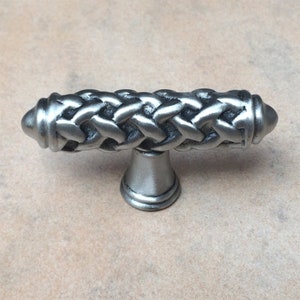 May include: A silver-toned cabinet pull with a braided design. The handle is horizontal with rounded ends and a decorative base. The hardware is made of metal and has a matte finish.