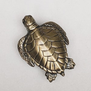 May include: A bronze-colored turtle figurine with detailed shell and flipper features. The sculpture has a textured surface and is designed to resemble a sea turtle. The figurine is a decorative item.