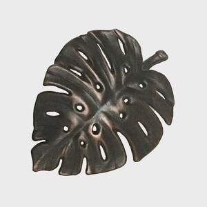 May include: A bronze-colored metal leaf with multiple holes and cutouts, resembling a Monstera plant leaf. The leaf has a textured surface and a stem. This decorative piece is suitable for wall decor or other home accents.