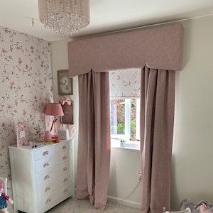 This picture shows a little girls bedroom with a pink and white coloured scheme.  

There is a large window with a partly closed roller blind.  The window also has a pink pelmet with a shaped bottom and matching floor length lined curtains.