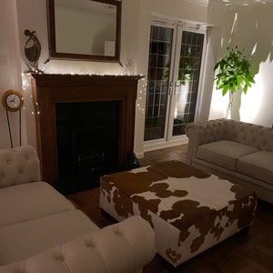 May include: A white couch with a brown and white cowhide ottoman in front of a fireplace with a string of lights. The room has a wooden floor and a large window with a view of a garden.