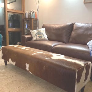 May include: A brown and white cowhide ottoman with wooden legs. The ottoman is in front of a brown leather sofa with a throw pillow featuring a bee design.