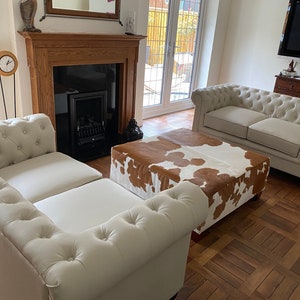 May include: A living room with two white leather sofas and a brown and white cowhide ottoman. The room has a fireplace with a black firebox and a wooden mantel. There is a mirror above the fireplace and a clock on the wall.