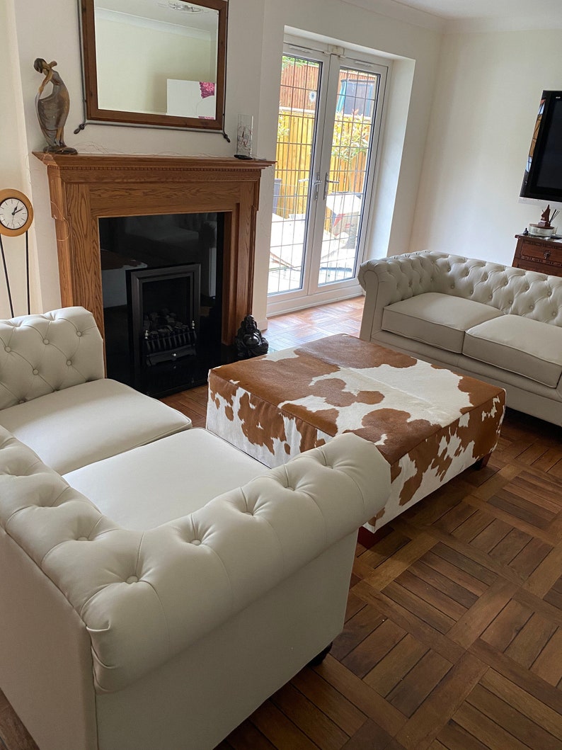 May include: A living room with two white leather sofas and a brown and white cowhide ottoman. The room has a fireplace with a black firebox and a wooden mantel. There is a mirror above the fireplace and a clock on the wall.