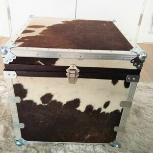 May include: A brown and white cowhide storage trunk with a silver metal frame and a silver metal latch. The trunk is sitting on a white fluffy rug.