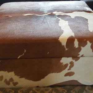 May include: A brown and white cowhide ottoman with a rectangular shape. The ottoman has a brown top and a white bottom with brown spots.
