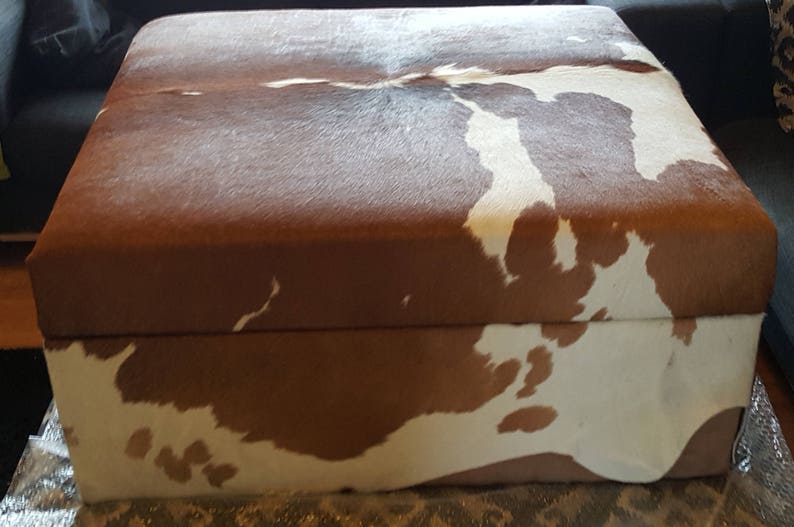May include: A brown and white cowhide ottoman with a rectangular shape. The ottoman has a brown top and a white bottom with brown spots.