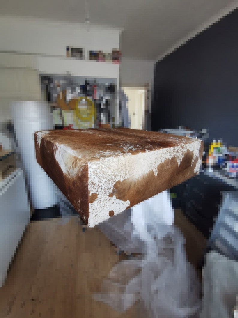 K&ouml;nnte beinhalten: Ein rechteckiger Hocker aus braunem und wei&szlig;em Kuhfell. Der Hocker steht auf einem Holzboden.