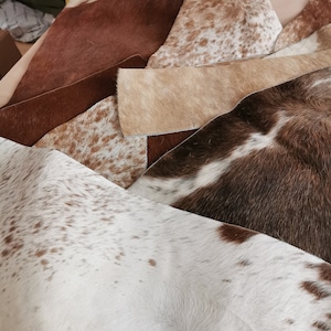 May include: A collection of assorted cowhide leather scraps featuring various natural patterns and textures. The pieces display a range of colors including solid brown, white with brown spots, tan, and dark brown with white patches in irregular shapes.