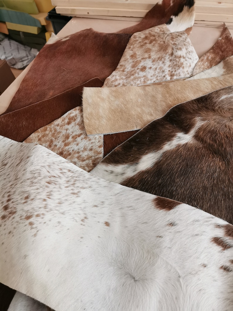 May include: A collection of assorted cowhide leather scraps featuring various natural patterns and textures. The pieces display a range of colors including solid brown, white with brown spots, tan, and dark brown with white patches in irregular shapes.