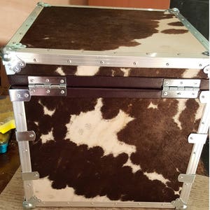 May include: A brown and white cowhide covered storage case with silver metal trim and hinges. The case has a rectangular shape and is made of sturdy material.