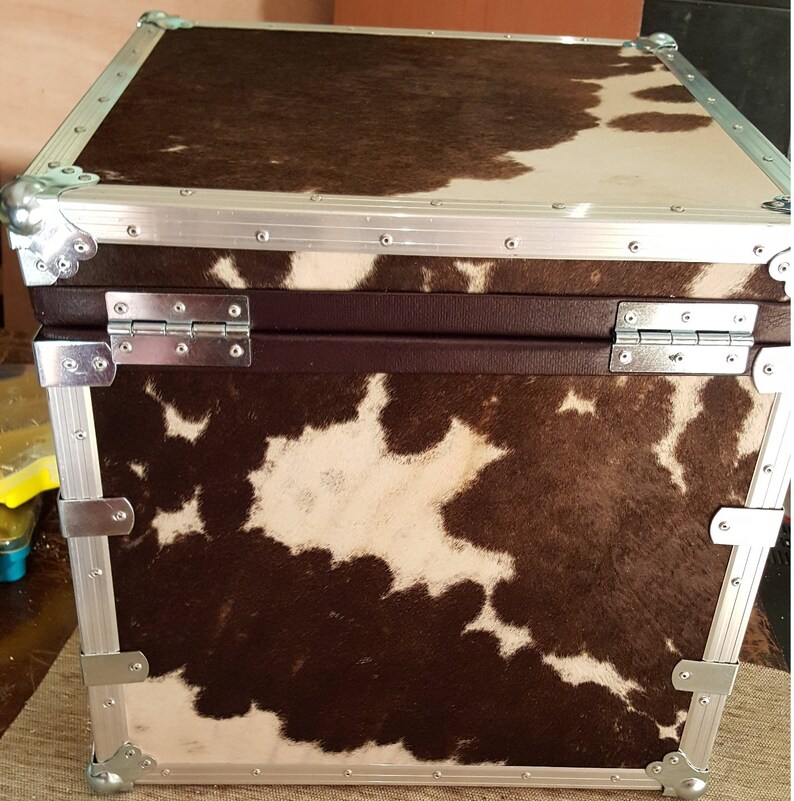 May include: A brown and white cowhide covered storage case with silver metal trim and hinges. The case has a rectangular shape and is made of sturdy material.