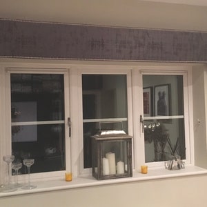 May include: A gray window valance with a white trim and a row of white beads along the edge. The valance is mounted above a white window frame with three panes of glass. The window is decorated with a lantern, candles, and other decorative items.