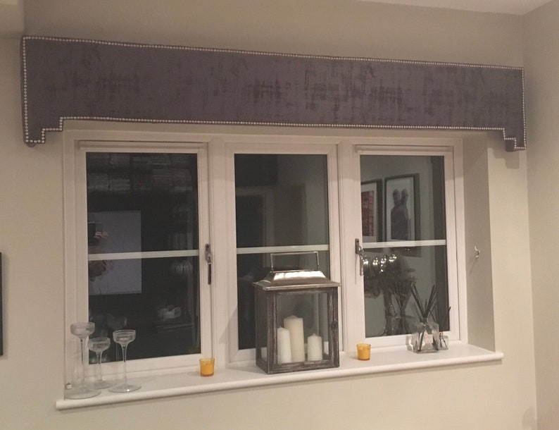 May include: A gray window valance with a white trim and a row of white beads along the edge. The valance is mounted above a white window frame with three panes of glass. The window is decorated with a lantern, candles, and other decorative items.