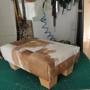 May include: A rectangular ottoman upholstered in a patchwork of brown and white cowhide, featuring a low-profile design with four light-colored wooden block feet. The surface displays a varied pattern of natural hair-on-hide textures.