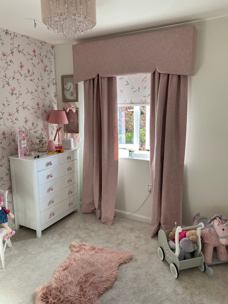 This picture shows a little girls bedroom with a pink and white coloured scheme.  

There is a large window with a partly closed roller blind.  The window also has a pink pelmet with a shaped bottom and matching floor length lined curtains.
