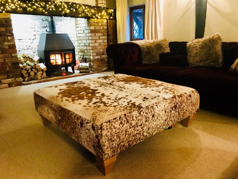 K&ouml;nnte beinhalten: Ein gro&szlig;er, quadratischer, brauner und wei&szlig;er Kuhfell-Ottomane mit einem braunen Holzrahmen. Der Ottomane befindet sich in einem Wohnzimmer mit einem Kamin und einer Couch im Hintergrund.