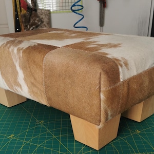 May include: A brown and white cowhide ottoman with wooden legs. The ottoman is sitting on a green cutting mat.