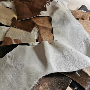 May include: A collection of assorted cowhide leather scraps featuring various natural patterns and textures. The pieces display a mix of colors including white, light gray, tan, dark brown, and black, with some sections showing spotted markings.