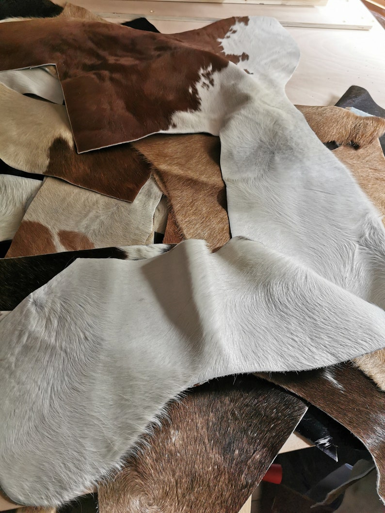 May include: A collection of assorted cowhide leather scraps featuring various natural patterns and textures. The pieces display a mix of colors including white, light gray, tan, dark brown, and black, with some sections showing spotted markings.