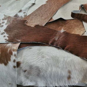 May include: A collection of overlapping cowhide leather scraps featuring various patterns and textures. The pieces display a mix of solid brown, solid white, and brown-and-white spotted fur, with visible raw edges and natural hair-on-hide surfaces.
