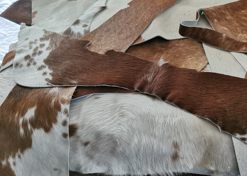 May include: A collection of overlapping cowhide leather scraps featuring various patterns and textures. The pieces display a mix of solid brown, solid white, and brown-and-white spotted fur, with visible raw edges and natural hair-on-hide surfaces.