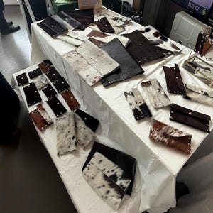 May include: A collection of rectangular leather and cowhide pouches and clutches arranged on a white surface. The items feature various textures, including solid dark brown leather and spotted white, black, and brown cowhide patterns with metal snaps.