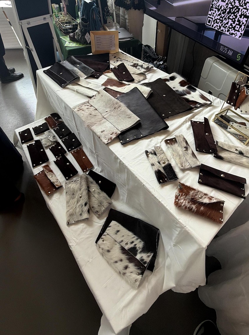 May include: A collection of rectangular leather and cowhide pouches and clutches arranged on a white surface. The items feature various textures, including solid dark brown leather and spotted white, black, and brown cowhide patterns with metal snaps.