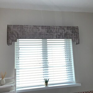 May include: A gray window valance with a textured pattern hangs above white blinds. A small potted plant sits on the windowsill.