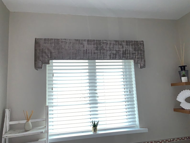May include: A gray window valance with a textured pattern hangs above white blinds. A small potted plant sits on the windowsill.