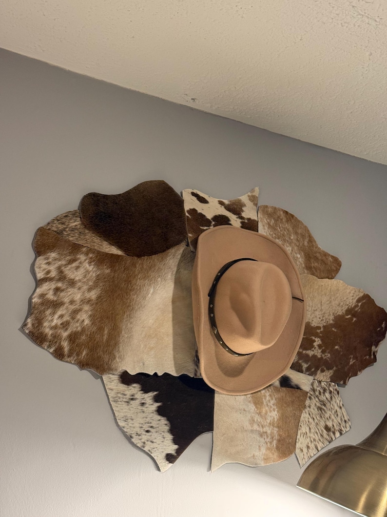May include: A tan cowboy hat with a dark band featuring metallic studs, mounted over a decorative wall display composed of multiple irregular patches of cowhide in shades of brown, white, and black, arranged in a layered, organic pattern.