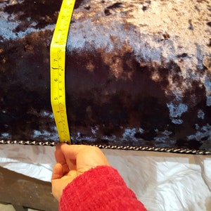 May include: Close-up of a dark blue velvet cushion with a silver and black pattern. A yellow measuring tape is being used to measure the length of the cushion, which is 17 inches.